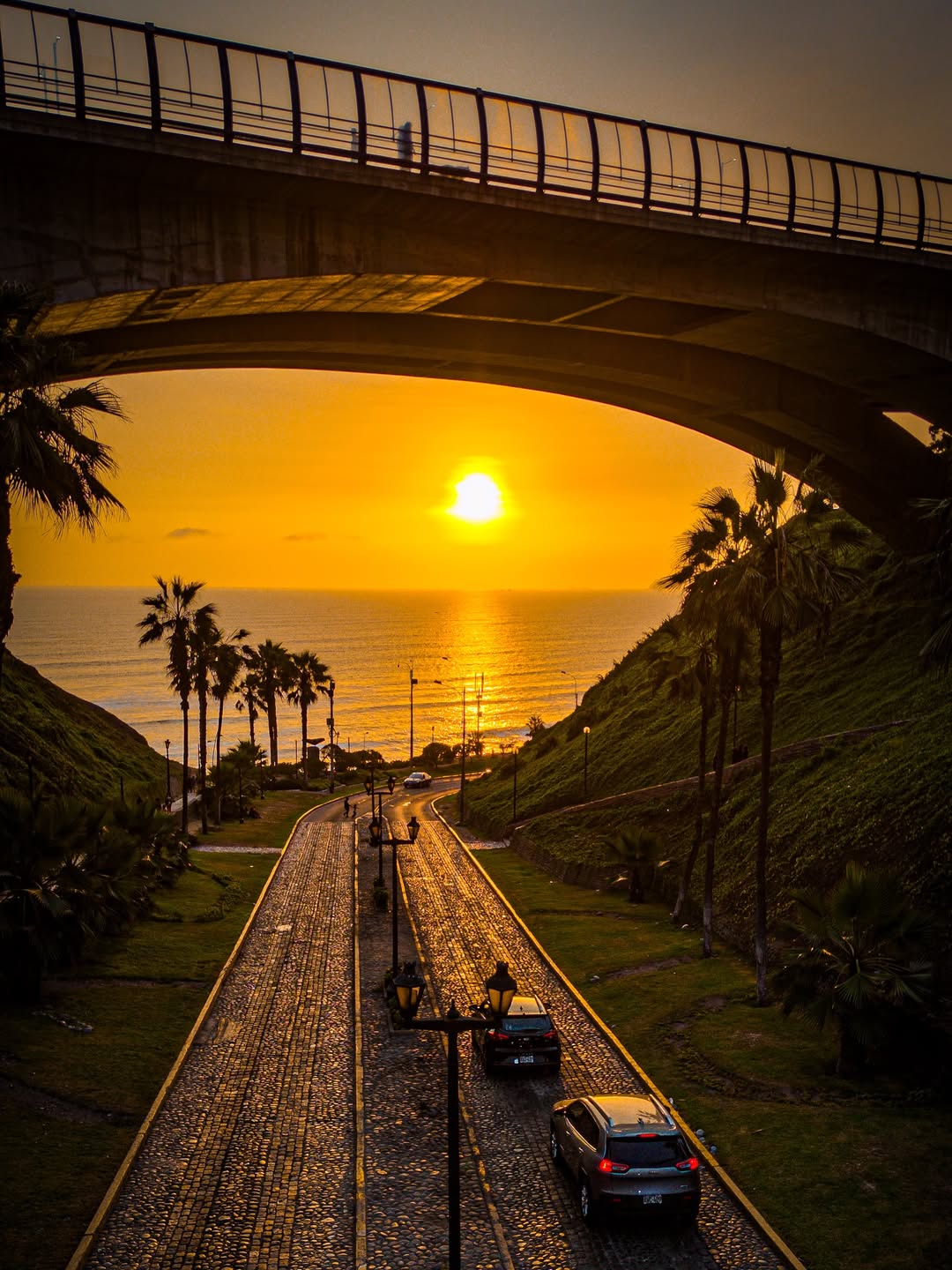 Coucher de soleil sur l’océan Pacifique vu depuis Miraflores à Lima avec une route pavée, des palmiers et un pont au-dessus.