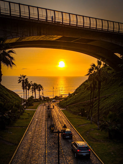 Coucher de soleil sur l’océan Pacifique vu depuis Miraflores à Lima avec une route pavée, des palmiers et un pont au-dessus.